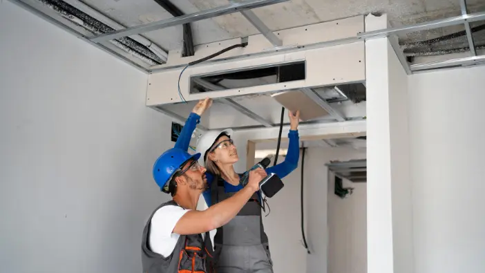 Two men collaborating to install or repair air ducts in a well-lit room, demonstrating teamwork and construction skills.