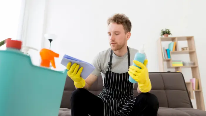 A man in rubber gloves sits on a couch, holding a cleaning product, ready to tackle household cleaning tasks.