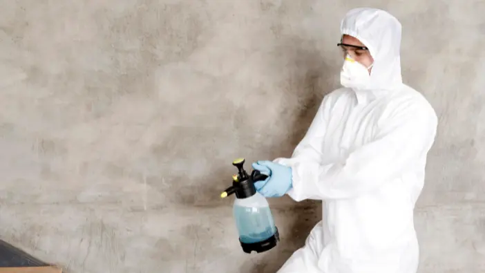 A man in a white suit and protective gear sprays a wall, ensuring safety and cleanliness in his work environment.