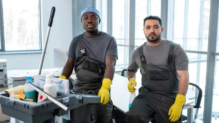 Two men in overalls and rubber gloves are actively cleaning a room, focusing on maintaining a clean and safe space.