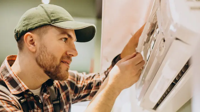 A man wearing a hat and plaid shirt cleans an air conditioner, focused on his task in a well-lit environment.